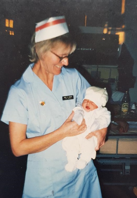 Photo of Christine Cooke at Southlands Hospital holding a baby, she is dressed in a pale blue nursing uniform.