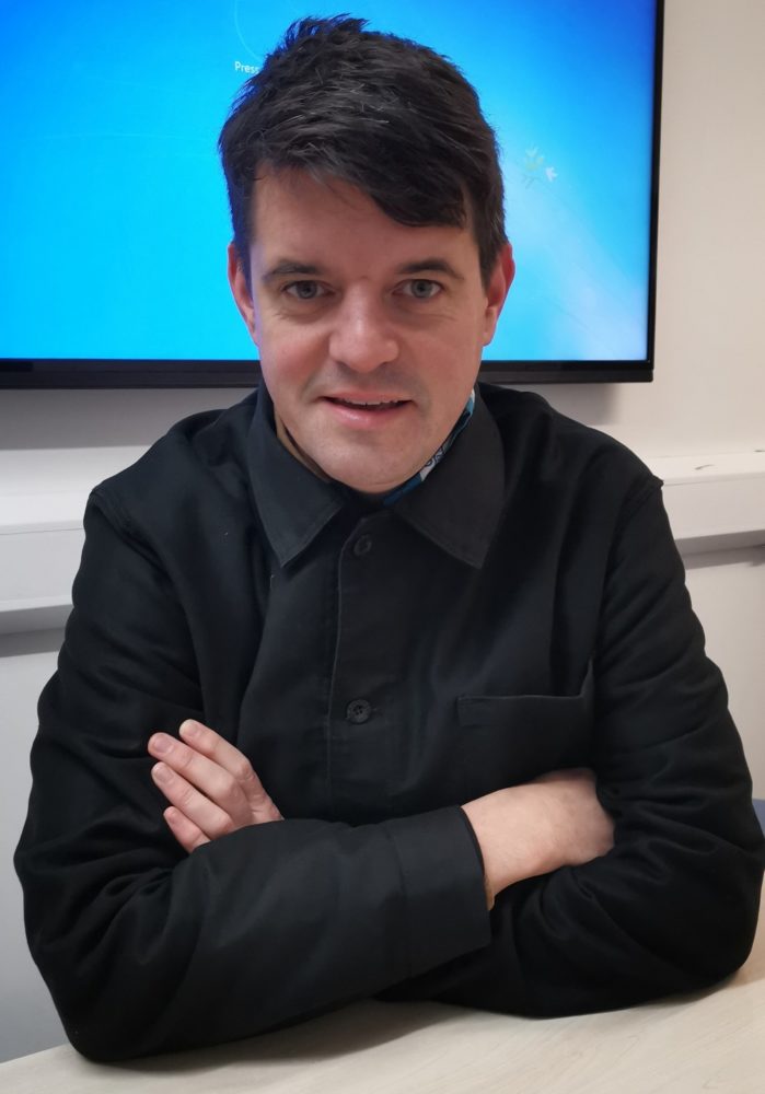 Photo of Daniel Locke sitting with arms crossed wearing a long-sleeved black shirt.